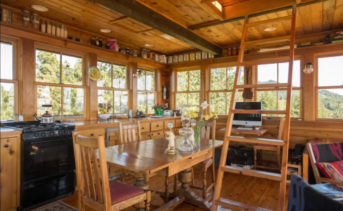 Cozy wooden interior of a cabin with a dining table, kitchen area, and a ladder leading to a loft. Bright windows offer scenic views.