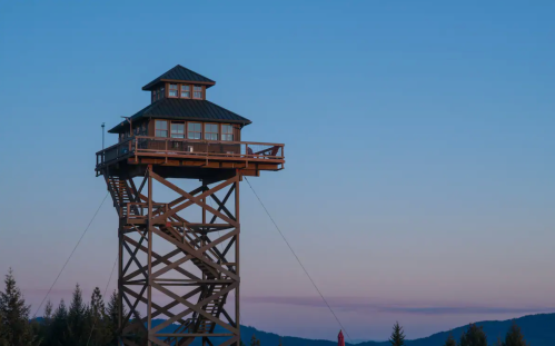 A tall wooden lookout tower with a peaked roof, set against a twilight sky and distant mountains.