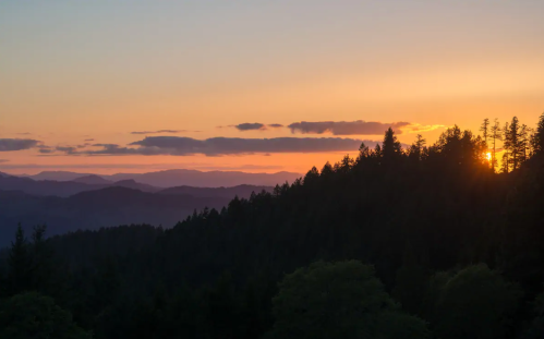 Sunset over a mountain range, with silhouettes of trees and layered hills in the background. Warm colors fill the sky.