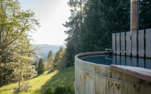 A wooden hot tub sits on a hillside, surrounded by trees and overlooking a scenic valley under a bright sky.