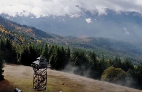 A wooden lookout tower stands in a misty valley surrounded by dense forests and mountains under a cloudy sky.