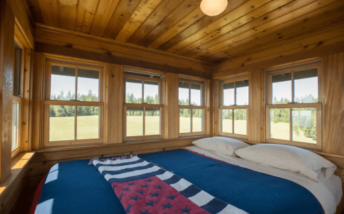 Cozy wooden bedroom with large windows, a blue bedspread, and a view of green fields outside.