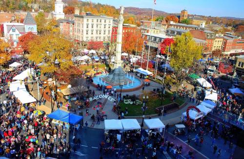 A bustling town square filled with people and colorful tents, surrounded by autumn foliage and historic buildings.
