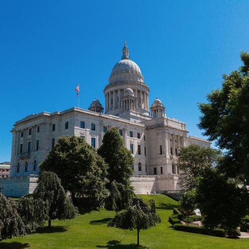 A grand stone building with a dome, surrounded by green trees and a clear blue sky.