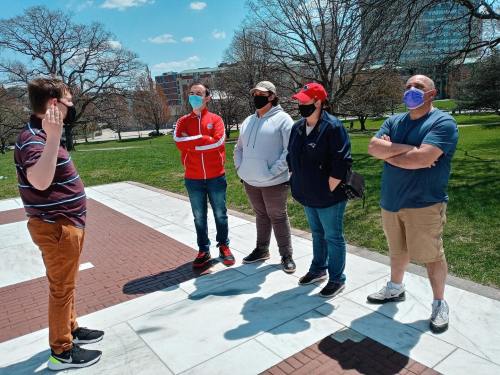 A group of five people wearing masks stands outdoors, engaged in conversation on a sunny day.