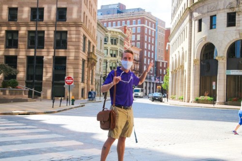 A person in a purple shirt and mask stands on a city street, waving with a bag over their shoulder.