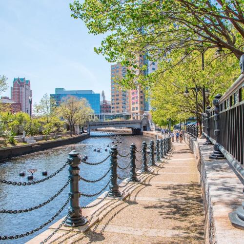 A scenic riverside path lined with trees and a chain fence, with buildings and a bridge in the background on a sunny day.
