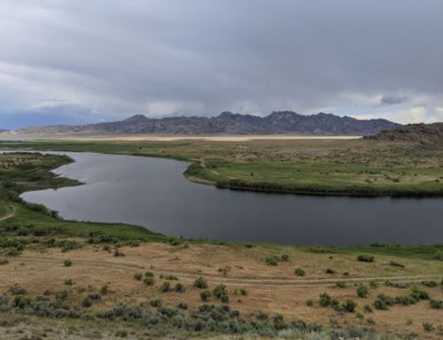 A serene river winding through green fields, with mountains in the background under a cloudy sky.