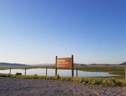 Sign for Pathfinder National Wildlife Refuge with a serene lake and distant mountains under a clear blue sky.