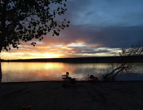 A serene lakeside scene at sunset, with vibrant colors reflecting on the water and a person fishing nearby.