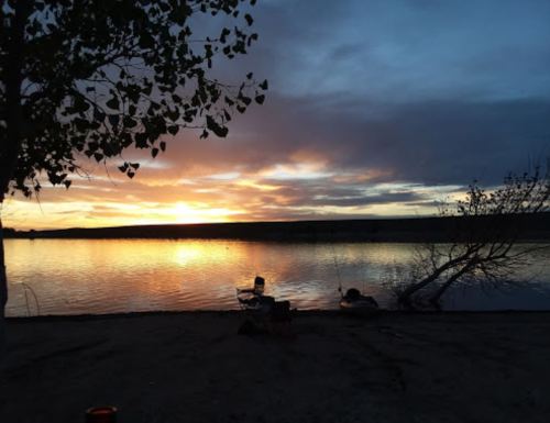 A serene lakeside scene at sunset, with vibrant colors reflecting on the water and a person fishing nearby.