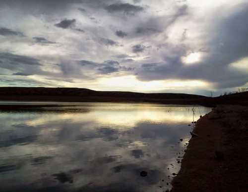 A serene lake at sunset, reflecting clouds and hills under a dramatic sky.