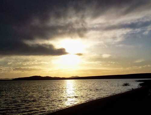 Sunset over a calm lake, with dark clouds and distant hills silhouetted against a golden sky.