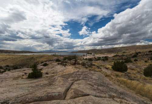 A rocky landscape with sparse vegetation under a cloudy sky, overlooking a lake in the distance.