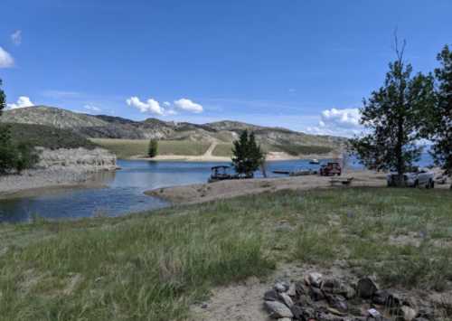 A serene lakeside view with grassy shores, distant hills, and a few boats on the water under a blue sky.