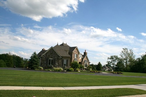 A large stone house with a manicured lawn under a blue sky with clouds, surrounded by trees and landscaping.