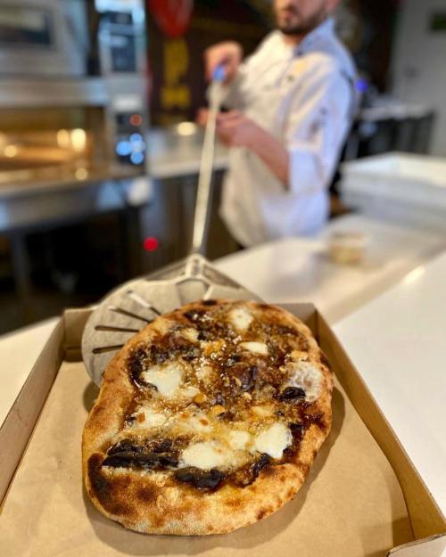 A freshly baked pizza with melted cheese, placed in a box, while a chef prepares to serve it in the background.