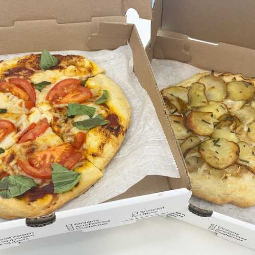 Two pizzas in a takeout box: one with tomatoes and basil, the other with sliced potatoes and herbs.