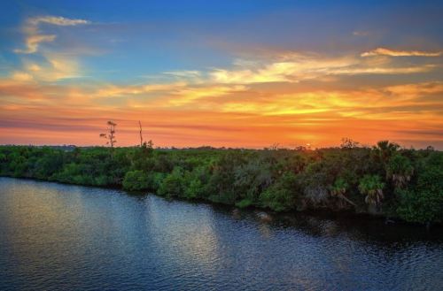A serene river scene at sunset, with vibrant skies reflecting on the water and lush greenery along the banks.
