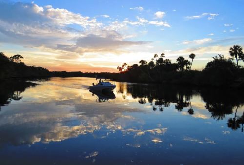 A boat glides across a calm river at sunset, reflecting vibrant clouds and palm trees in the water.