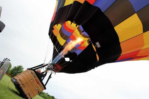 A colorful hot air balloon inflates as flames shoot from the burner, with a wicker basket below on green grass.