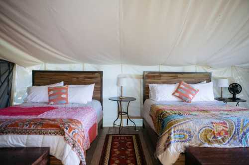 Cozy glamping tent interior with two beds, colorful blankets, and a small table with a lamp.