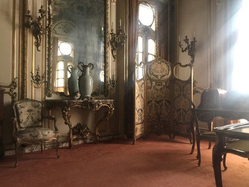 Elegant room with ornate furniture, a large mirror, and soft light filtering through windows, creating a vintage atmosphere.