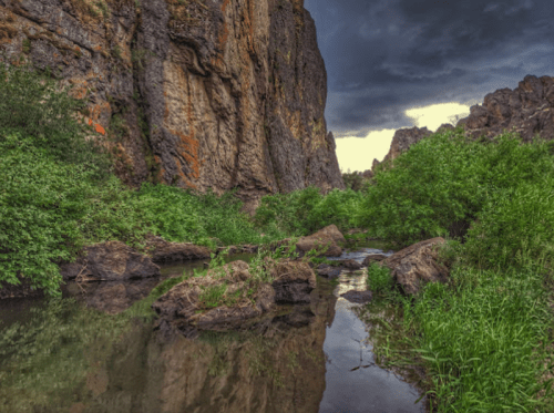 A serene landscape featuring a rocky canyon, lush greenery, and a calm stream under a cloudy sky.