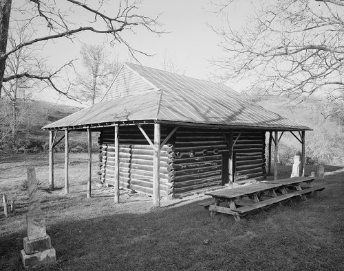 A rustic log cabin with a metal roof, surrounded by trees and a picnic table in a grassy area. Black and white photo.