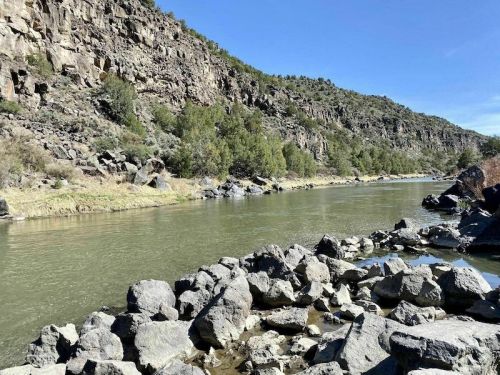 A serene river flows between rocky banks, surrounded by lush greenery and steep cliffs under a clear blue sky.
