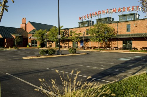 Exterior view of a brick building with "Riverton Market" sign, surrounded by trees and an empty parking lot.
