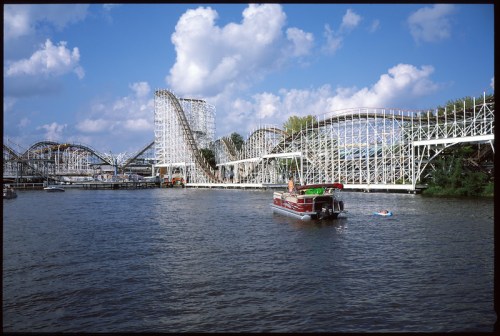 A scenic view of a roller coaster by a river, with a boat and a person swimming nearby under a blue sky.
