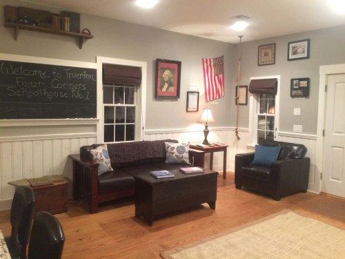 Cozy schoolhouse interior with a chalkboard, American flag, and seating area featuring a couch and armchair.