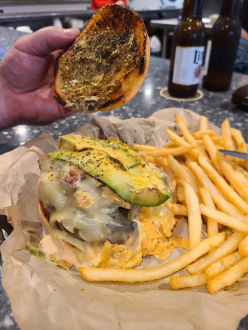 A hand holds a toasted burger bun above a loaded cheeseburger with avocado, served with fries on the side.