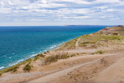 A sandy dune overlooks a vibrant blue ocean under a cloudy sky, with distant cliffs visible along the shoreline.