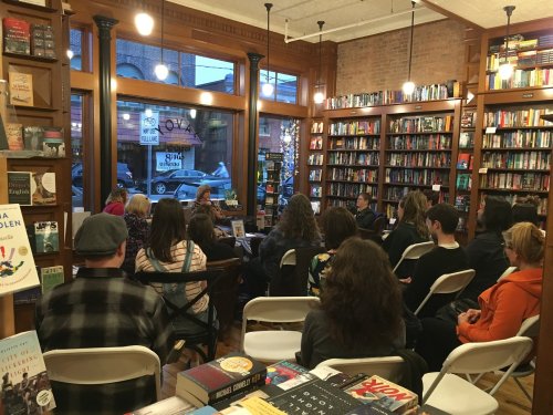 A cozy bookstore event with an audience seated, listening to speakers among shelves of books and warm lighting.