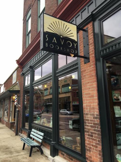 Exterior of Savoy Bookshop with a sign, large windows, and a bench outside on a brick building.