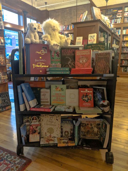 A wooden book cart filled with holiday-themed books and plush toys in a cozy bookstore setting.