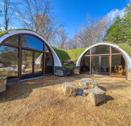 Two modern, curved buildings with green roofs, surrounded by trees and a gravel area with large rocks in the foreground.