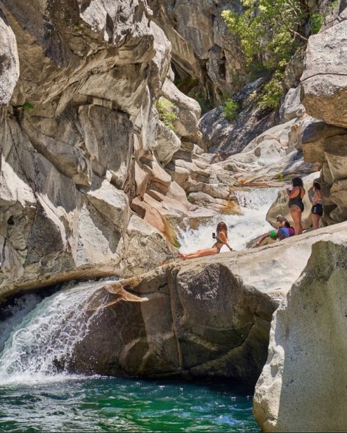 A group of people relaxes by a rocky river with waterfalls and lush greenery in a sunny outdoor setting.
