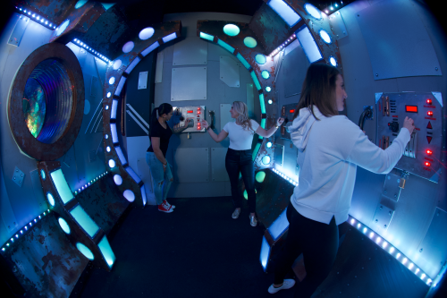 Three people interact with control panels in a futuristic, illuminated room resembling a spaceship or sci-fi setting.