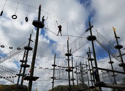 A high ropes course with people navigating obstacles among tall wooden structures against a cloudy sky.