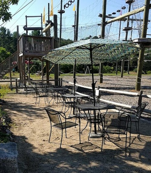 Outdoor seating area with tables and chairs under an umbrella, near a ropes course and playground.
