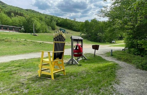 A large yellow chair and a red bell stand on a grassy path, surrounded by trees and a cloudy sky.