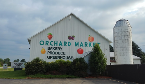 A white barn with "Orchard Market" painted on the side, featuring images of fruits and a silo nearby.