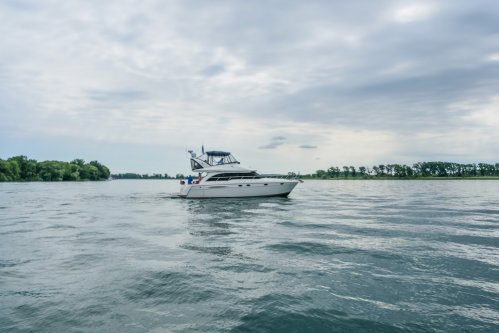 A sleek white yacht glides across calm waters under a cloudy sky, surrounded by lush greenery along the shore.
