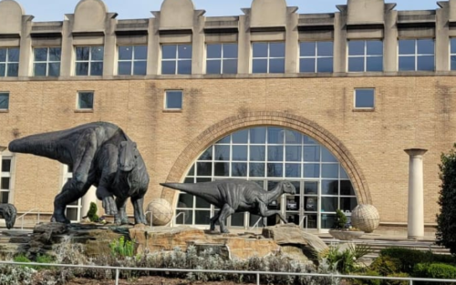 Two large dinosaur sculptures in front of a brick building with large windows and a curved entrance.