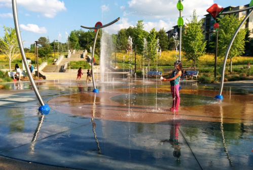 Children play in a splash pad, enjoying water features and a sunny day in a park setting.