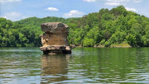A large rock formation stands in calm water, surrounded by lush green trees and a clear blue sky.