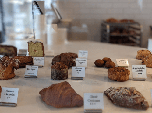 A display of various baked goods, including cookies, muffins, and pastries, with price tags in a bakery setting.